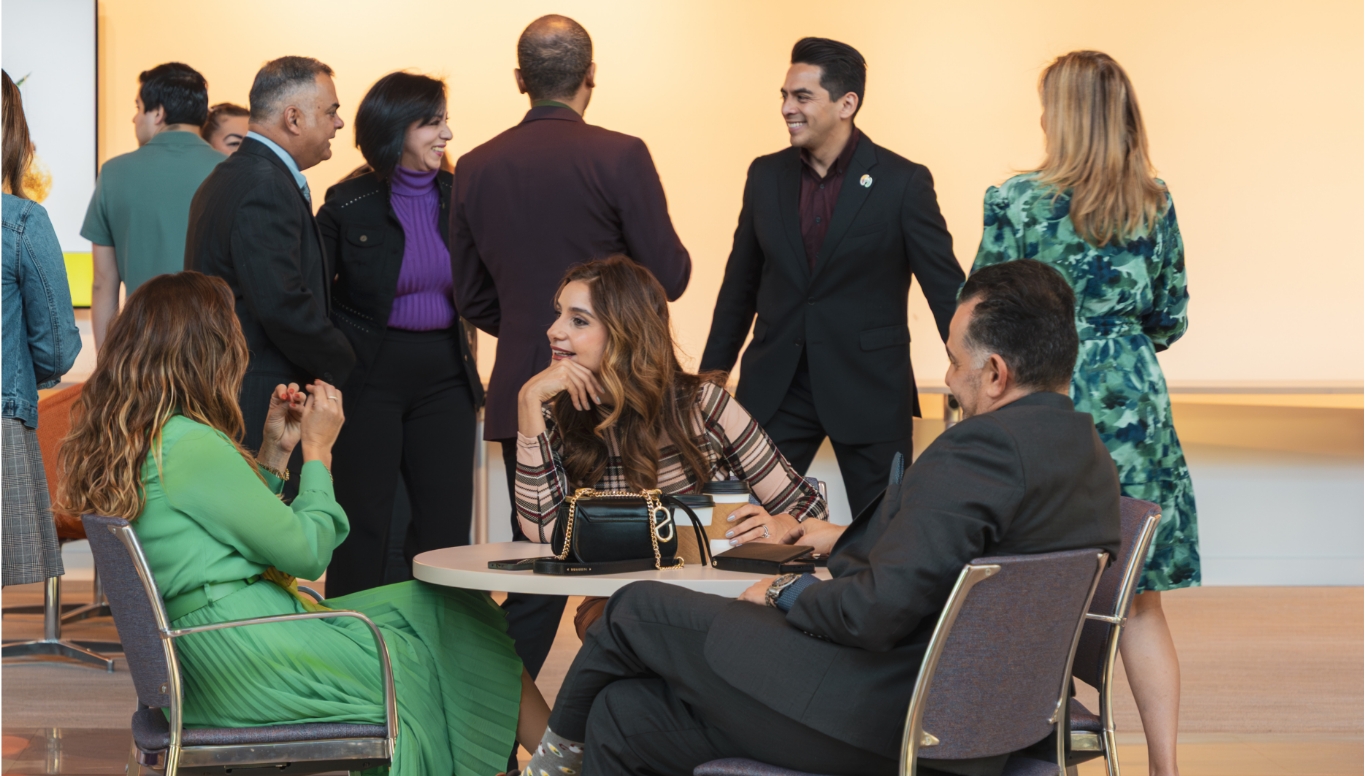 A group of people, dressed in semi-formal attire, sit and stand in a brightly lit room, smiling and chatting with each other about Winter Park Health Foundation. Three people are seated at a round table in the foreground.