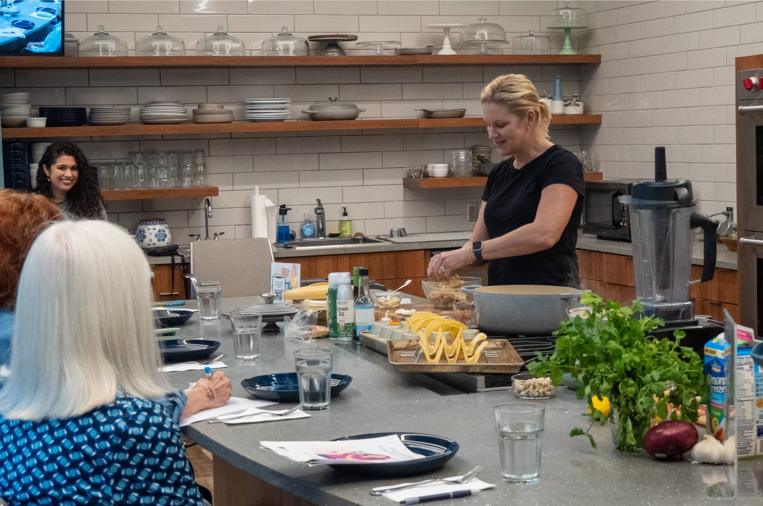A woman is giving a cooking demonstration in a modern kitchen while several people watch from a counter. The counter displays fresh ingredients, utensils, and glasses of water. For more information, contact Winter Park Health Foundation. Shelves line the background.